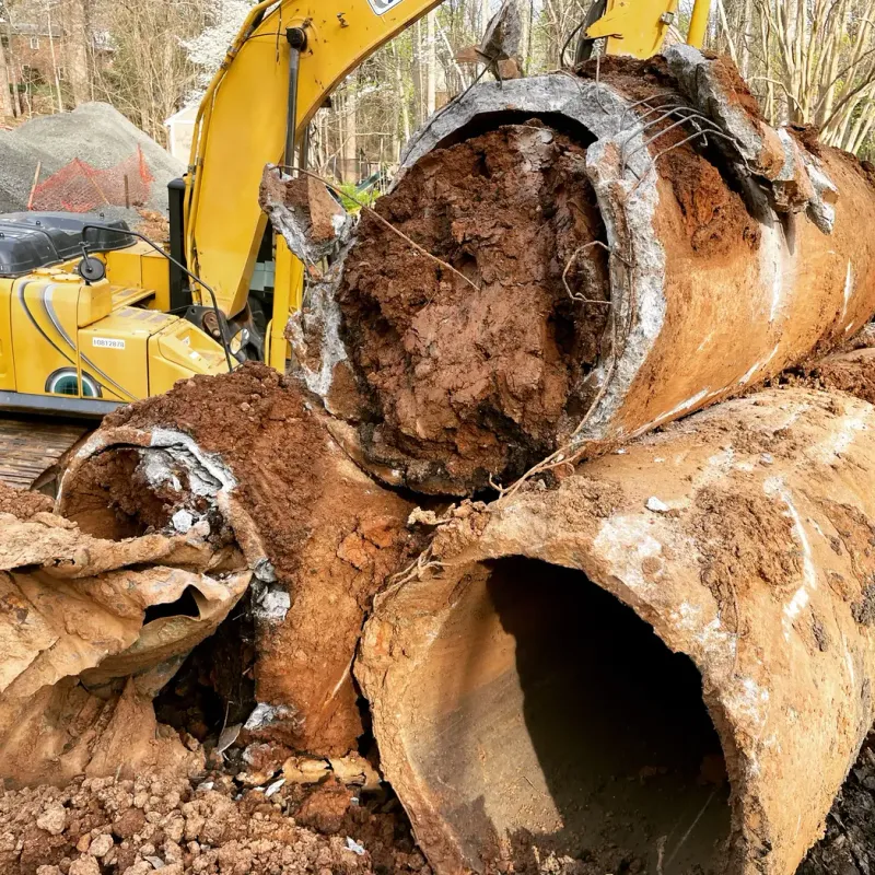 A large, broken concrete pipe lies in a muddy construction site, with a yellow excavator in the background.
