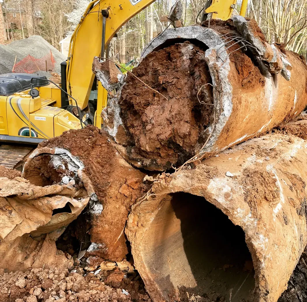 A large, broken concrete pipe lies in a muddy construction site, with a yellow excavator in the background.